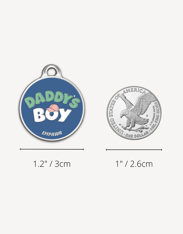 Empawr "Daddy's Boy" charm next to a US dollar coin for size comparison. The charm is 1.2 inches (3 cm) in diameter, and the coin is 1 inch (2.6 cm). The charm is blue with white letters and a pink baseball cap graphic.