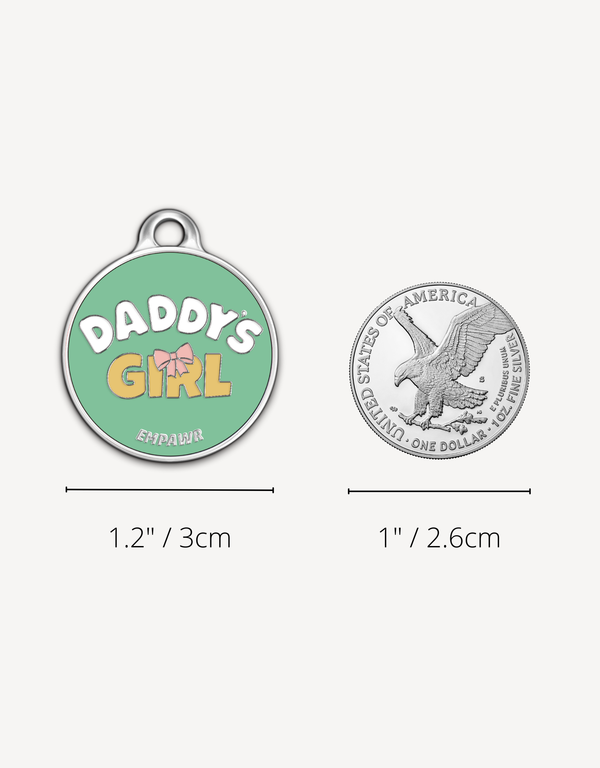 Empawr "Daddy's Girl" charm next to a US dollar coin for size comparison. The charm is 1.2 inches (3 cm) in diameter, and the coin is 1 inch (2.6 cm). The charm is green with yellow letters and a pink bow.
