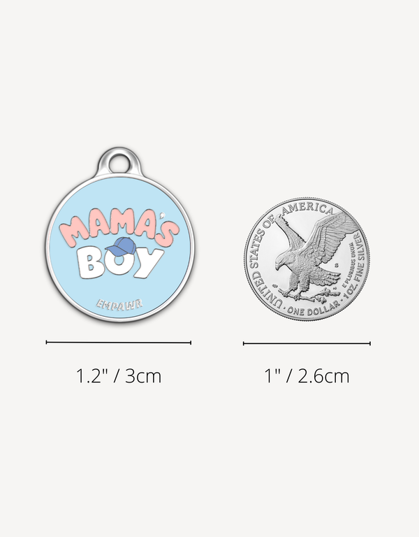 Empawr "Mama's Boy" charm next to a US dollar coin for size comparison. The charm is 1.2 inches (3 cm) in diameter, and the coin is 1 inch (2.6 cm). The charm is light blue with pink letters and a blue baseball cap graphic.