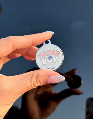 A hand with manicured nails holds a round charm that reads "Mama's Boy" in pink letters on a light blue background. The charm has a silver rim and a small loop at the top. The hand is held against a blurred background with a dark, reflective surface.